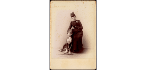 Studio portrait of unidentified woman in black dress and monocle with cigarette posing with Great Dane; whip and glove on floor